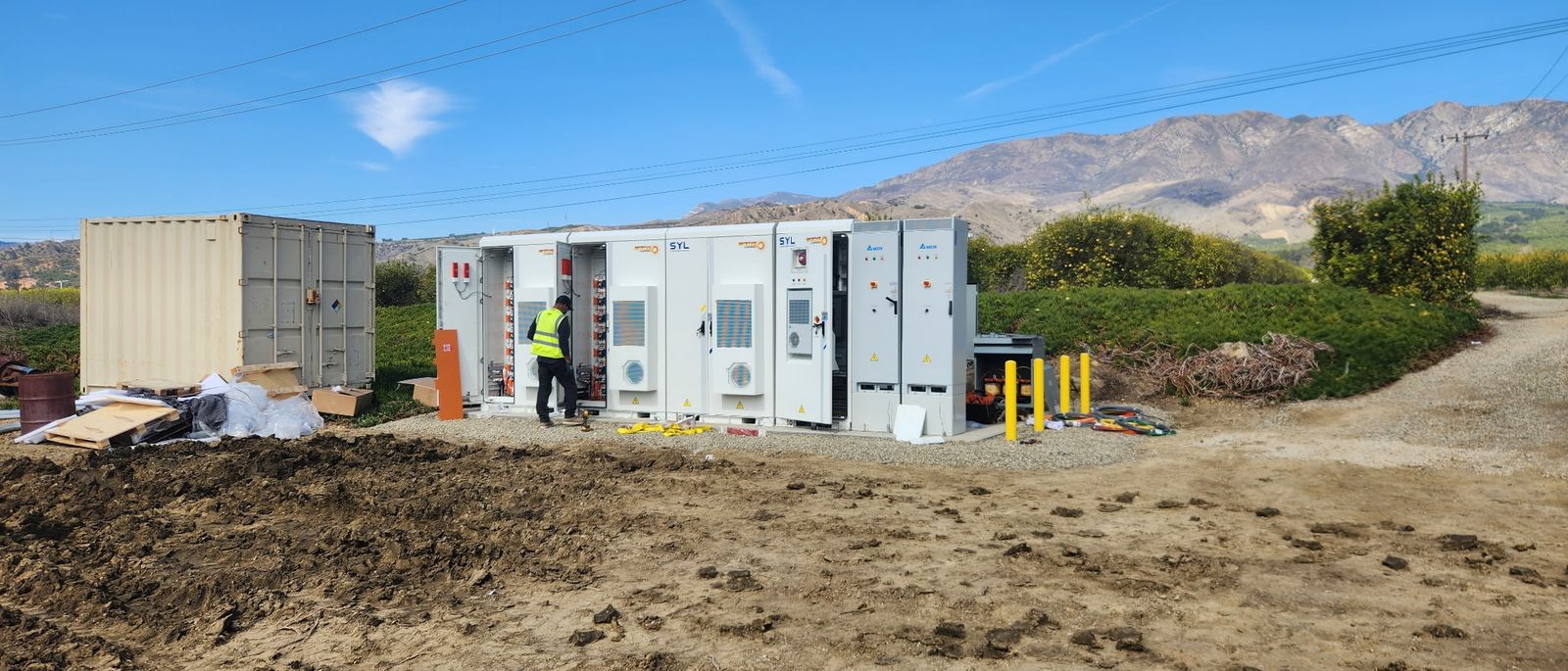 A technician in high-visibility clothing working at a row of battery cabinets, mountains behind