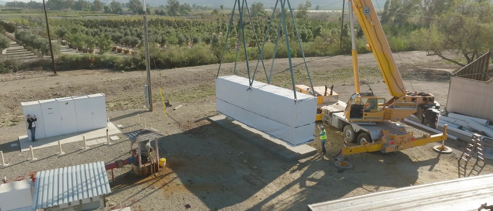 A large battery enclosure suspended from a crane on spreader bars above a prepared pad