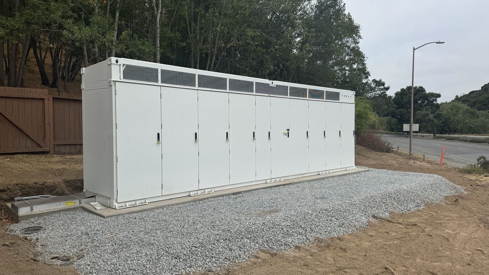 A long white battery enclosure on a fresh gravel pad beside a road, trees behind