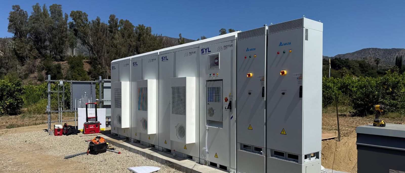 A row of white battery and inverter cabinets on gravel, orchard and hills behind